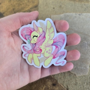 May include: A yellow and pink cartoon pony with a pink mane and tail, wearing a green bow. The pony is flying with its wings spread out. The sticker is on a white background.