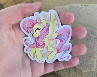 Könnte beinhalten: Ein gelbes und rosa Cartoon-Pony mit rosa Mähne und Schweif, das eine grüne Schleife trägt. Das Pony fliegt mit ausgebreiteten Flügeln. Der Aufkleber ist auf weißem Hintergrund.