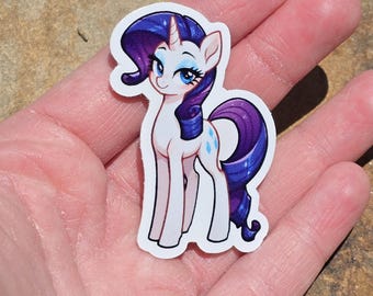 May include: A sticker of a white pony with a purple mane and tail, a blue diamond on its flank, and a unicorn horn. The pony has blue eyes and is smiling. The sticker is on a white background.