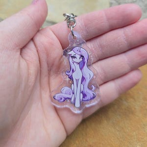 Puede incluir: Un llavero de acrílico transparente con un poni de dibujos animados con largo cabello morado y un cuerno de unicornio. El poni está representado sentado con una expresión triste. El llavero tiene un anillo de metal plateado.
