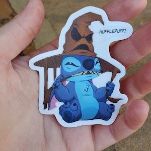 Peut inclure: Un personnage bleu Stitch portant un chapeau de tri de Poudlard brun avec le texte "HUFFLEPUFF!" sur le côté. Le personnage est assis sur une chaise avec une baguette magique dans sa main.