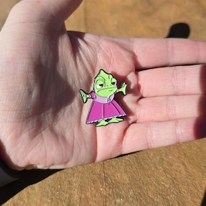 May include: A green chameleon wearing a pink dress with a white collar. The chameleon is standing on a hand with its arms outstretched.