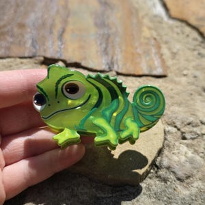 May include: A green chameleon-shaped brooch with a glossy finish. The brooch features a detailed design with various shades of green and yellow, including stripes and a curled tail. The chameleon has large, expressive eyes and is resting on a stone surface.