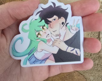 Puede incluir: Una pegatina con una ilustración de estilo anime de una pareja. La mujer tiene el pelo largo verde y ojos amarillos, y el hombre tiene el pelo negro y una camisa negra. Se abrazan y se miran.