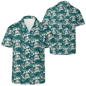 Puede incluir: Camisa de manga corta con fondo verde azulado y un estampado repetido de cascos de fútbol americano con diseño de cara de lobo. La camisa tiene cuello en V y un corte holgado, adecuada para ropa informal.