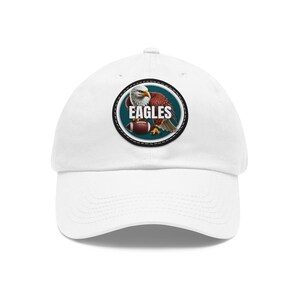 May include: White baseball cap featuring a circular patch with an eagle holding a football. The patch has the word "EAGLES" in white text. The eagle is depicted with brown and white feathers, and the football is brown and white.