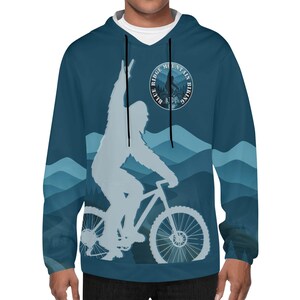 Puede incluir: Sudadera con capucha azul con una silueta blanca de un Bigfoot en bicicleta de montaña. El diseño incluye una cordillera y el texto "BLUE RIDGE MOUNTAIN BIKING". La sudadera tiene una capucha con cordón y un bolsillo delantero.