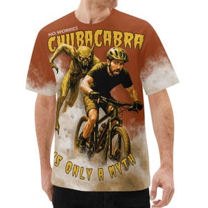 Puede incluir: Camiseta marrón con un diseño gráfico de un ciclista perseguido por un Chupacabra. El texto "No Worries Chupacabra It's Only a Myth" está en amarillo y naranja. La camiseta tiene cuello redondo y mangas cortas.