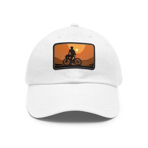 Peut inclure: Casquette de baseball blanche avec un écusson rectangulaire représentant un cycliste à contre-jour devant un coucher de soleil. L'écusson est bordé de noir. Le design évoque les activités de plein air et le cyclisme.