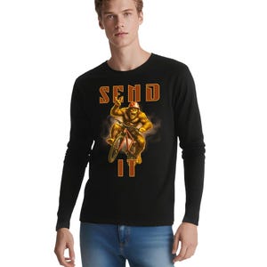 Pode incluir: Camiseta preta de manga comprida com um gráfico de um Bigfoot andando de bicicleta de montanha. O texto "SEND IT" é exibido em letras de bloco laranja acima e abaixo da ilustração do Bigfoot. O Bigfoot está usando um capacete vermelho e fazendo o sinal de paz.