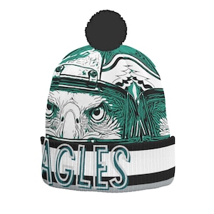 May include: A knit beanie featuring a detailed graphic of an eagle's face in teal and white. The word "EAGLES" is displayed in teal and white block letters on the brim. The hat has a black pom-pom on top and a black and gray stripe.