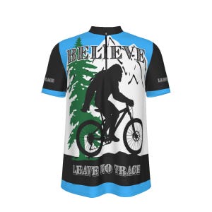 Pode incluir: Uma camisola de ciclismo de manga curta com um design azul, preto e branco. A frente apresenta uma silhueta de um Bigfoot a andar de bicicleta de montanha, com a palavra "BELIEVE" acima e "LEAVE NO TRACE" abaixo.