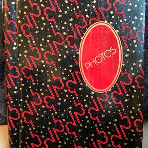 May include: A black photo album with a red and gold design. The cover features a repeating pattern of red stylized numbers and hearts, with small gold dots. A red oval label with the word "PHOTOS" in gold lettering is centered on the cover.
