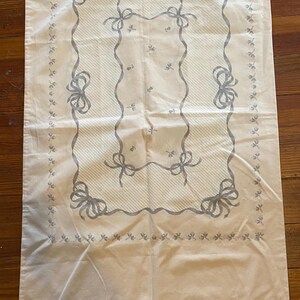 May include: A pale pink rectangular textile with a gray bow and ribbon design. The design is centered and framed by a border of small gray floral accents. The textile is likely a tablecloth or decorative fabric.