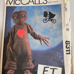 May include: A McCall's sewing pattern envelope for an E.T. The Extra-Terrestrial costume, featuring a brown creature with a red heart on its chest, a silhouette of a person on a bicycle against a full moon, and the pattern number 8311 printed on it.