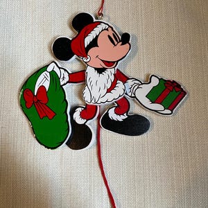 May include: A wooden Christmas ornament featuring Mickey Mouse dressed as Santa Claus. Mickey is wearing a red Santa hat and suit with white trim, holding a green stocking with a red bow and a wrapped gift. A red string is attached.