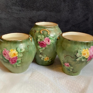 May include: Three antique green vases with gold rims and handles. Each vase is adorned with hand-painted pink and yellow roses and green leaves. The vases are of varying sizes and shapes, creating a decorative set.