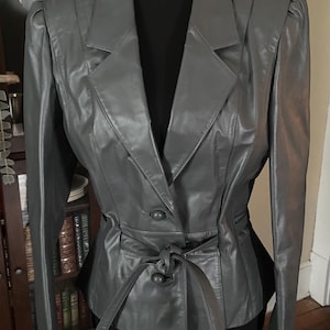 May include: A gray leather jacket with a notched collar, two-button closure, and a tie belt. The jacket has shoulder pads and a fitted silhouette. The jacket is displayed on a black mannequin.