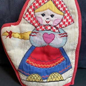 May include: A quilted oven mitt featuring an embroidered character with yellow hair, a red and white houndstooth headscarf, a blue skirt, and yellow shoes. The character holds a pink heart against a cream-colored background with a red border.