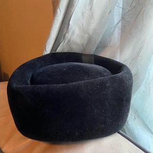May include: A solid black pillbox hat with a smooth, felt-like texture. The hat features a rounded, structured crown with a flat top and a wide, upright brim. A small, dark ribbon detail is visible on the side of the crown, adding a subtle accent to the otherwise uniform, deep black surface of the headwear.