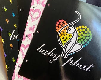 May include: A stack of Baby Phat branded folders. The folders have a black background with a white cat logo and the words "baby phat" in cursive. Other folders have heart and cat patterns.