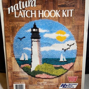 May include: A "natura LATCH HOOK KIT" featuring a Cape Cod design. The kit includes a round rug with a lighthouse, sailboats, and birds against a blue sky and yellow sun. The rug is approximately 23 inches in diameter.