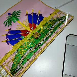 May include: A pink embroidered square with the Haitian coat of arms, featuring a palm tree, flags, and the motto "L'Union Fait La Force." A yellow ruler, marked in inches, is placed diagonally across the fabric, measuring approximately 12 inches.
