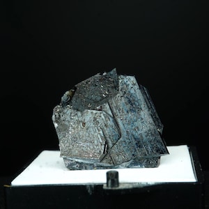 Puede incluir: Un espécimen mineral oscuro y metálico con una estructura geométrica compleja. La superficie muestra una mezcla de tonos plateados, grises y azules, con una apariencia cristalina. El mineral se encuentra sobre una superficie blanca sobre un fondo negro.
