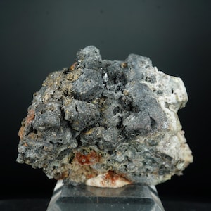 Puede incluir: Primer plano de un espécimen mineral con una superficie rugosa y texturizada. La roca presenta una mezcla de colores gris oscuro, negro y gris claro, con toques de marrón rojizo. El mineral se encuentra sobre un soporte transparente sobre un fondo negro.