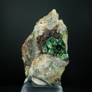 May include: A rough-hewn rock formation with a cavity of vibrant green mineral deposits. The exterior displays a mix of beige, brown, and gray tones, creating a natural, textured appearance. The mineral specimen is displayed on a clear stand against a dark background.