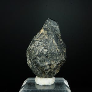 Puede incluir: Un espécimen mineral de color gris oscuro, de forma irregular, posiblemente un meteorito, exhibido sobre un soporte transparente con una base blanca. La roca tiene una superficie rugosa y texturizada con parches más claros.