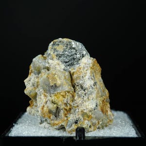May include: A mineral specimen with a rough, textured surface. The rock displays a mix of beige, grey, and dark grey metallic sections. It rests on a bed of small, white crystalline fragments against a black backdrop. The mineral's structure is uneven and complex.