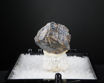 May include: A dark blue and brown mineral specimen, possibly a crystal, sits atop a white, porous base within a black display case. The mineral has a rough, textured surface and is illuminated against a dark background.