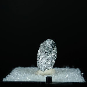 Op de afbeelding: Een zilverkleurig mineraalmonster, mogelijk een metaalerts, wordt tentoongesteld op een bed van helder, gemalen glas in een zwarte vitrine. Het mineraal heeft een ruw, getextureerd oppervlak en is gemonteerd op een kleine, gebroken witte basis.