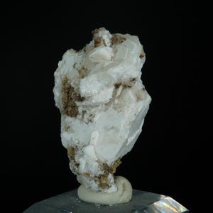 May include: A white mineral specimen with a layered, crystalline structure. The mineral has a rough, textured surface with patches of brown and tan inclusions. The specimen is mounted on a small, rounded, off-white base against a black background.