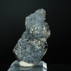 Può includere: Un campione di minerale grigio scuro con una superficie ruvida e strutturata. Il minerale presenta piccole inclusioni scintillanti di colore dorato ed è montato su una base rettangolare trasparente. Lo sfondo è nero.