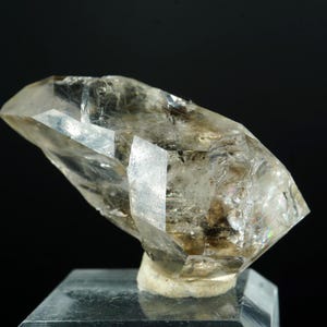 May include: A clear quartz crystal with multiple facets, resting on a dark surface. The crystal has a light brown hue with internal inclusions and is illuminated against a black background. This natural mineral specimen is a unique decorative item.
