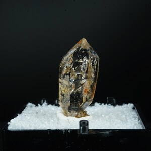 May include: A clear, faceted crystal with amber and brown hues. The crystal is displayed on a white base within a black square frame, set against a dark background. The crystal's surface reflects light, showcasing its natural structure.