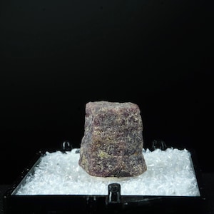 May include: A rough-cut, rectangular ruby gemstone with a dark purple hue. The ruby is displayed on a black platform filled with small, clear, crystalline pieces. The background is a solid black.