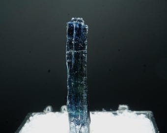 May include: A vertically oriented, dark blue crystal with a rough, textured surface. The crystal is displayed in a black tray filled with white material, set against a dark gray background. The crystal's color transitions to a lighter blue and green in some areas.