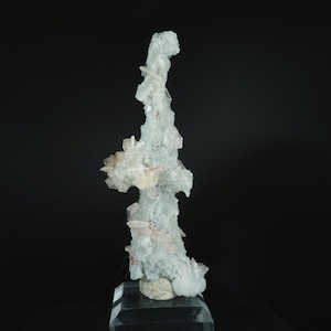 May include: A tall, vertical mineral specimen featuring a cluster of pale, translucent, and white crystalline structures. The formation has a rough, textured surface with protruding, sharp-edged crystal points and sits on a clear square base.