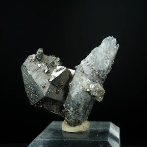 May include: A mineral specimen with a cluster of metallic grey and crystalline formations. The specimen has a dark, geometric shape on the left and a translucent, light-coloured crystal structure on the right. The mineral sits on a small, light-coloured base.