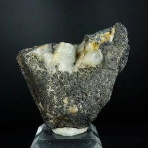 May include: A rough, dark gray rock formation with crystalline white and yellow mineral deposits. The specimen is displayed on a clear, angled stand against a black background, highlighting its natural texture and color variations.