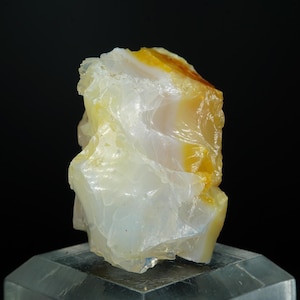 Puede incluir: Una gema translúcida en bruto con una mezcla de tonos blancos, amarillos y anaranjados. La piedra se exhibe sobre un pedestal claro e inclinado contra un fondo negro, resaltando su textura natural y variaciones de color.