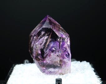 May include: A faceted amethyst crystal with a deep purple hue and clear sections. The crystal is displayed on a white, textured surface against a black background. The crystal's pointed tip and unique internal patterns are visible.