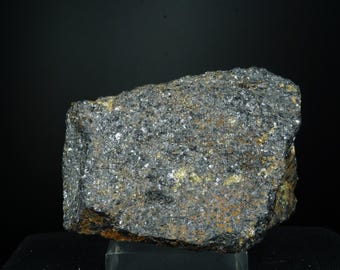 Può includere: Una formazione rocciosa ruvida, di forma irregolare, con un aspetto grigio scuro e argentato metallico. La superficie presenta una struttura ruvida e cristallina con sfumature dorate e marroni. La roccia è esposta su sfondo nero.