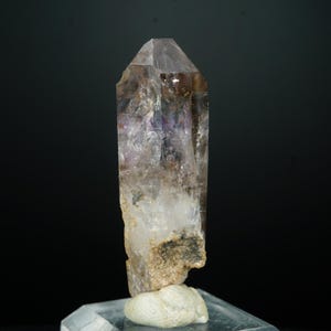 May include: A clear quartz crystal with a pointed tip. The crystal has a gradient of colors, including purple, brown, and white. The base of the crystal is a rough, beige color. The crystal is displayed on a clear, angled surface against a black background.