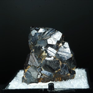 May include: A dark, metallic mineral specimen with a cluster of sharp, geometric crystal formations. The crystals have a reflective surface, with shades of black and silver. The mineral sits on a bed of white, crystalline material.