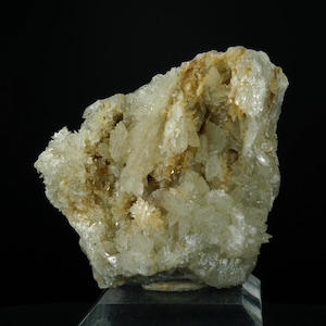 Può includere: Un campione di minerale cristallino di colore chiaro con una superficie ruvida e strutturata. Il minerale presenta un mix di aree traslucide e opache, con sfumature di marrone e giallo. Il campione è esposto su sfondo nero.