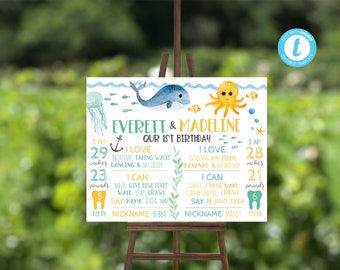 May include: A blue and yellow first birthday milestone sign with a whale and octopus illustration. The sign includes the names Everett and Madeline, their ages in inches and pounds, and things they love and can do.